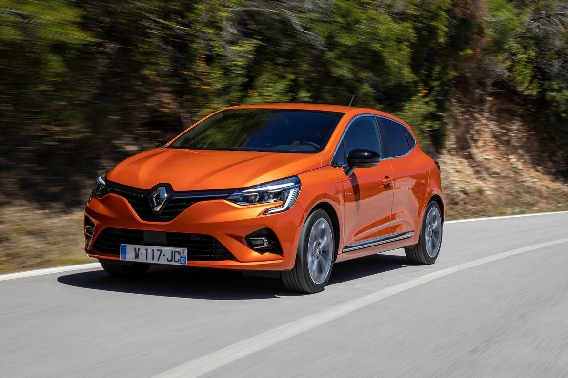 Układ jezdny Renault Clio V udanie łączy komfort oraz dobre prowadzenie Pomarańczowy samochód Renault Clio jedzie po asfaltowej drodze otoczonej drzewami i krzewami.