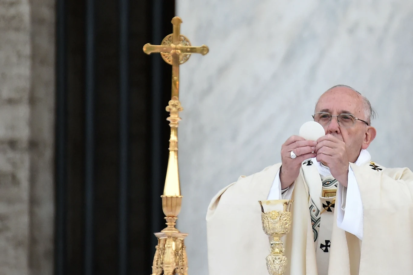 Ksiądz celebruje mszę świętą, unosząc hostię nad złotym kielichem. Obok znajduje się złoty krzyż na ołtarzu w tle z marmurową ścianą.