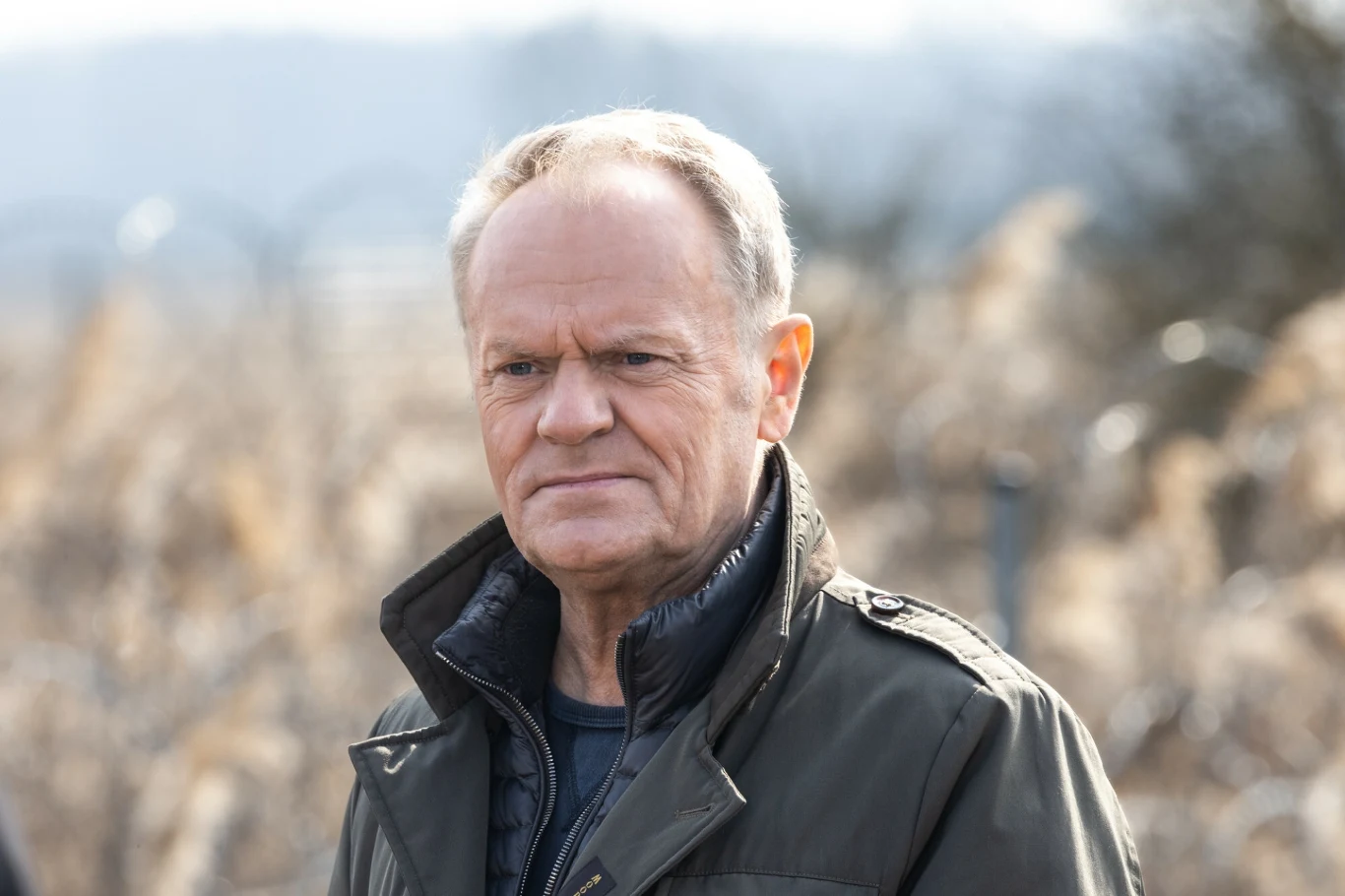 Donald Tusk jedzie do Biebrzańskiego Parku Narodowego Starszy mężczyzna z krótko ostrzyżonymi włosami nosi ciemną kurtkę, stoi w naturalnym otoczeniu na tle nieostrego pejzażu.