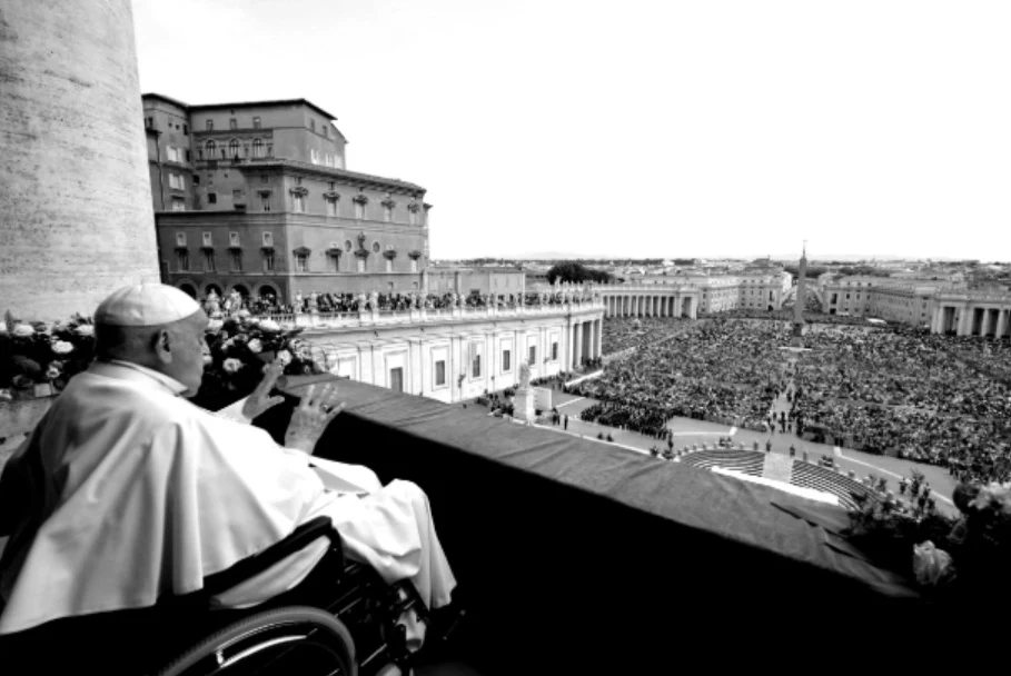 Nowe informacje ws. Franciszka. Watykan o ważnym terminie Papież w białym stroju siedzi na wózku inwalidzkim na balkonie, błogosławiąc zgromadzone na placu tłumy. Na pierwszym planie dostrzegalny jest balkon i kwiaty, w tle widoczny rozległy plac wypełniony wiernymi.