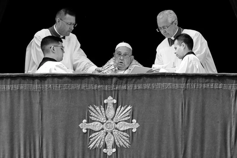 Papież Franciszek nie żyje. Dzień wcześniej zaskoczył wiernych Papież w otoczeniu czterech duchownych czytających dokument. Centralnym punktem jest papież, a zdjęcie utrzymane jest w czarno-białej tonacji. Przed nimi znajduje się ozdobny krzyż na obrusie.