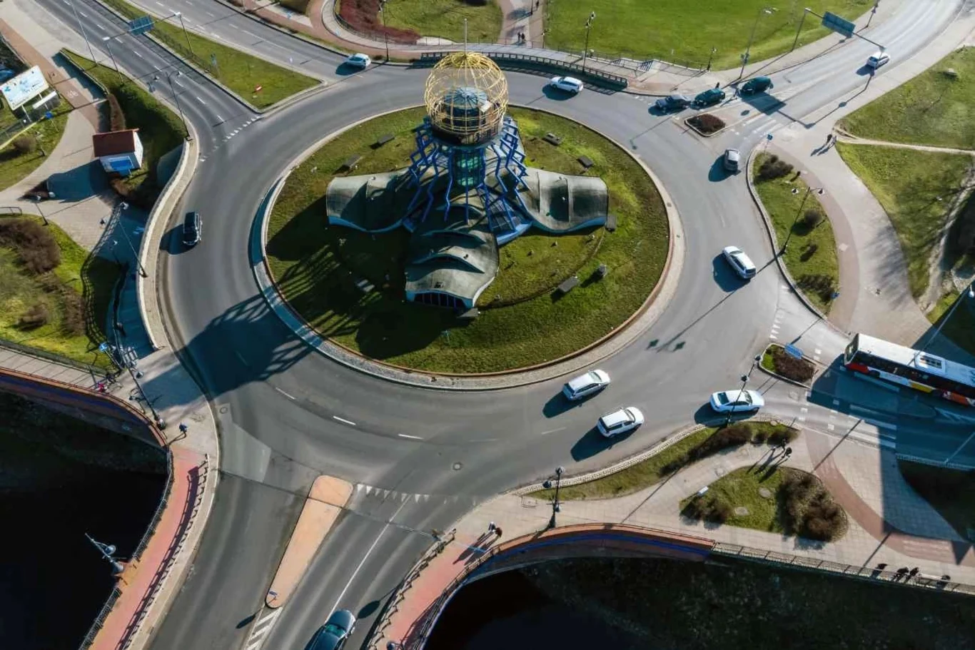 Na rondzie ze znakiem A-7 panują inne zasady dotyczące pierwszeństwa niż te, do których przywykli kierowcy Widok z lotu ptaka na rondo z charakterystycznym budynkiem o nowoczesnym kształcie w centrum, otoczonym przez zieloną przestrzeń.