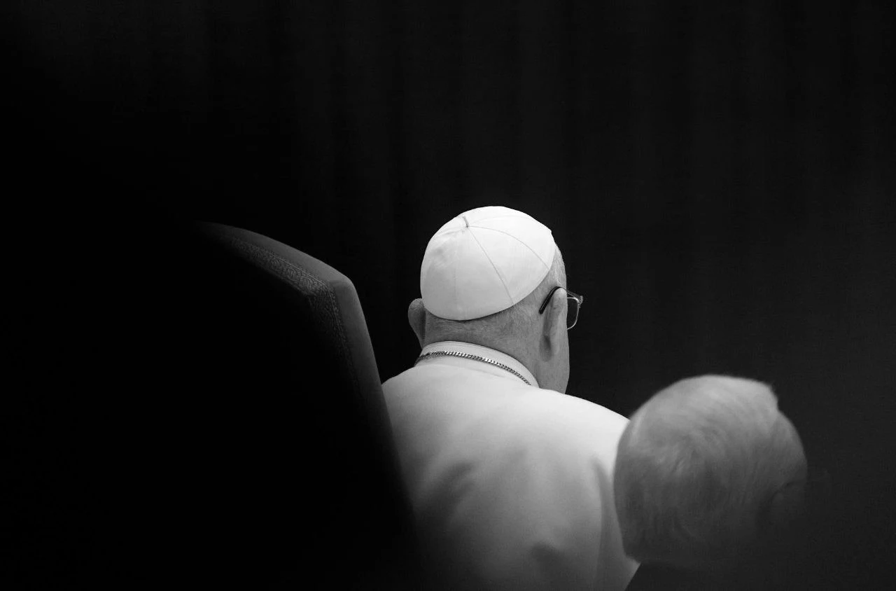Papież Franciszek nie żyje. Watykan opublikował przyczynę śmierci Ojca Świętego oraz jego testament Postać w białej sutannie i piusce siedząca w ciemnym pomieszczeniu widziana od tyłu, przyciąga uwagę minimalistyczna kompozycja i kontrast między białą szatą a otoczeniem.