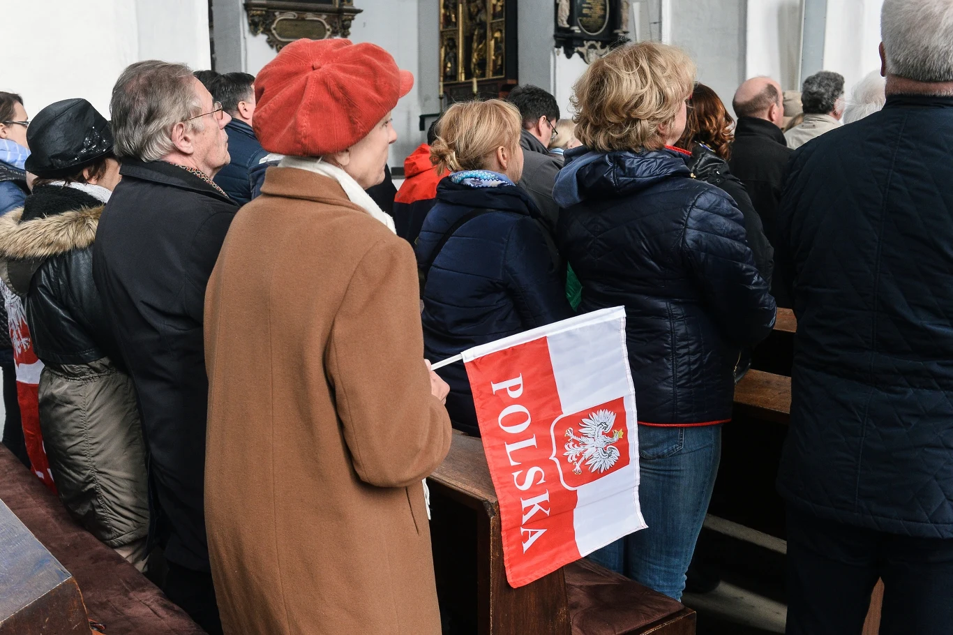 Uroczystość Najświętszej Maryi Panny Królowej Polski w Gdańsku Grupa osób zgromadzona wewnątrz budynku, jedna kobieta w czerwonym berecie trzyma flagę Polski. Ludzie zwróceni są w jednym kierunku, pomieszczenie ma jasne ściany i drewniane elementy wystroju.