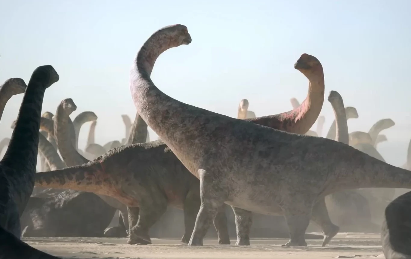 Największy północnoamerykański dinozaur. Odkryto szczątki sprzed 70 mln lat. Grupa dinozaurów na prehistorycznym krajobrazie, w otoczeniu kurzu i światła dziennego, tworzy dynamiczną scenę. W centrum uwagę przykuwa duży dinozaur z długą szyją.