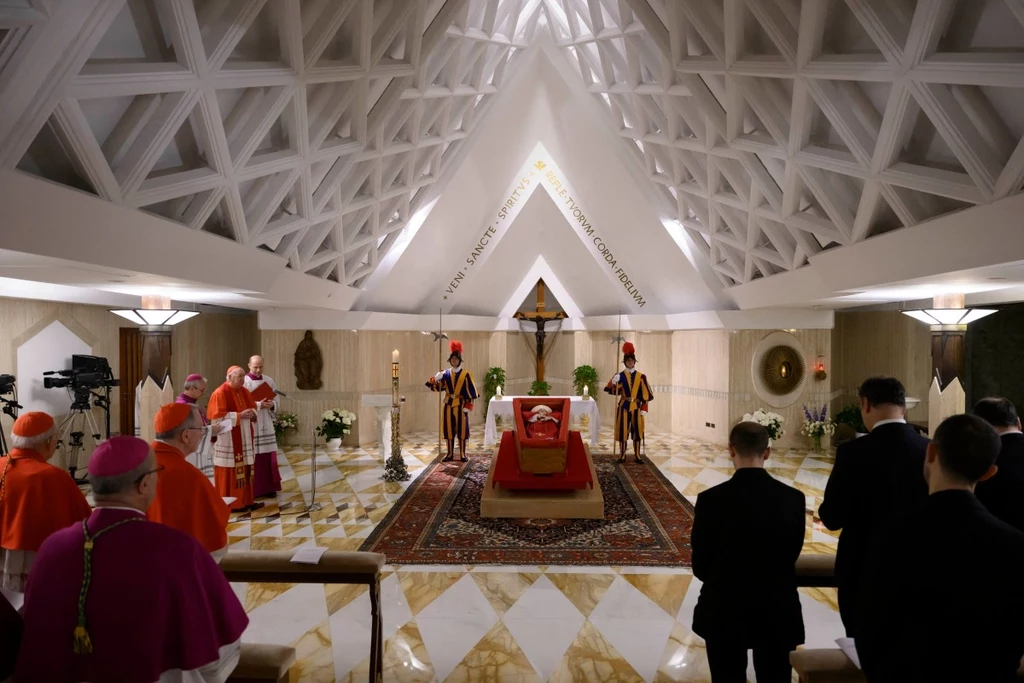 W kaplicy Domu Świętej Marty wystawiono trumnę z ciałem papieża Franciszka Wnętrze kaplicy z wysokim sklepieniem i trumną umieszczoną na czerwonym dywanie. Po obu stronach stoją gwardziści, a wokół zgromadzeni są kardynałowie i inni uczestnicy w czarnych strojach. Ściany są ozdobione krzyżem i rozetami.
