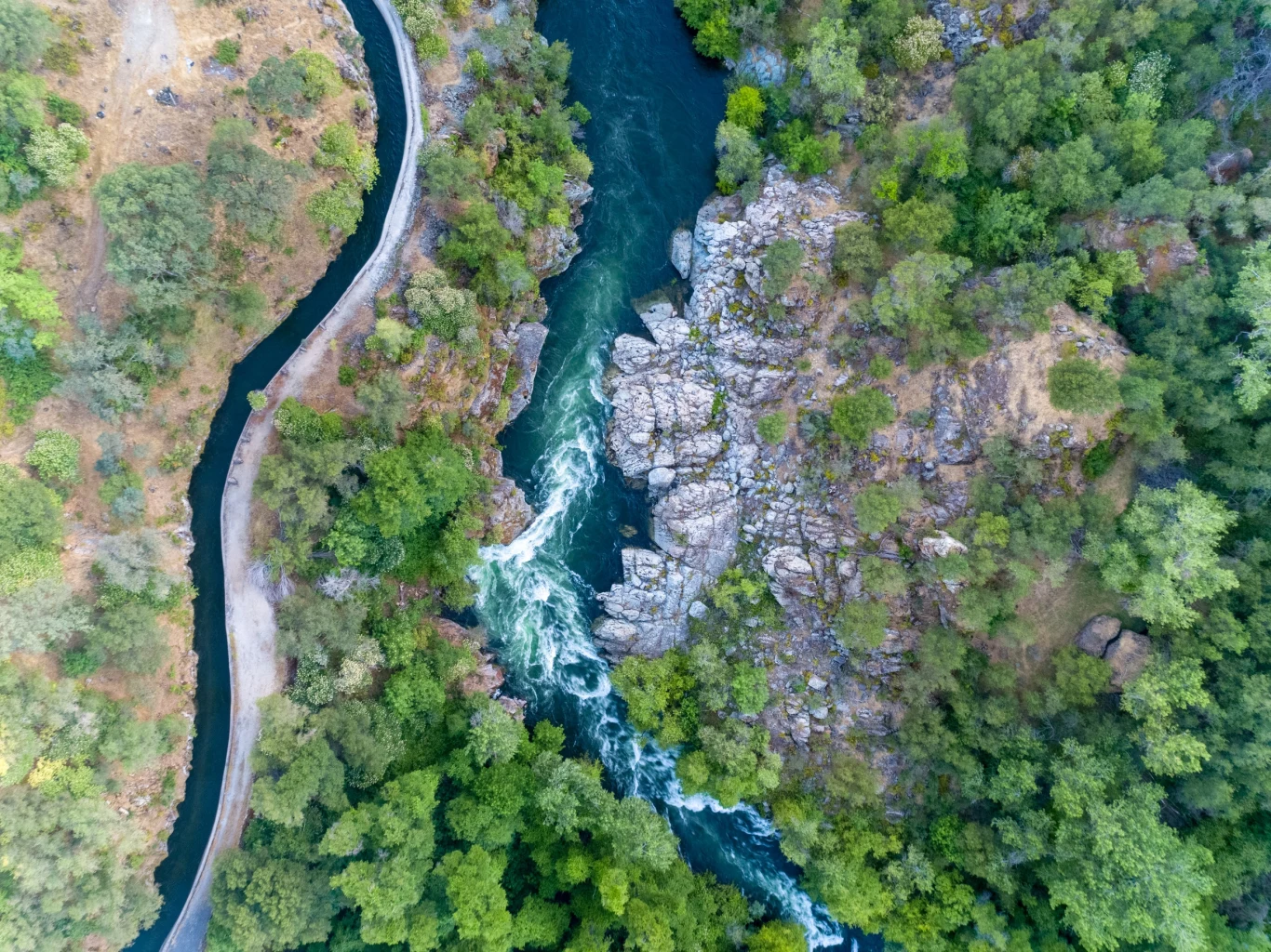 Widok z lotu ptaka na rzekę przepływającą przez gęsty las z licznymi drzewami. Rzeka tworzy zatoczki i wodospady, wijąc się przez zielony krajobraz. Na lewo od rzeki widać wąską drogę.