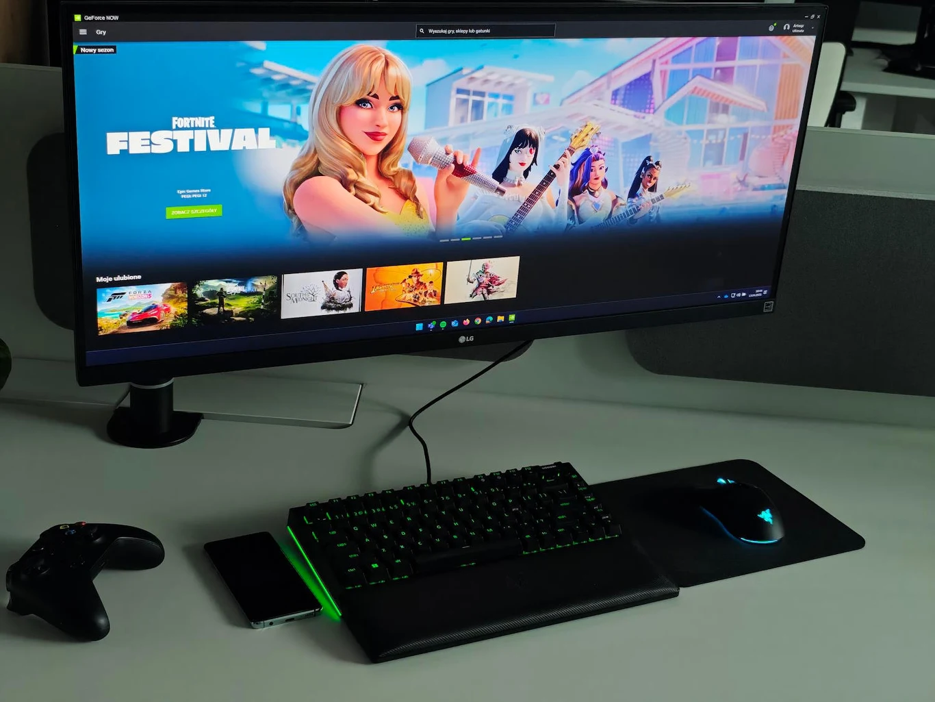 Stanowisko do gier z dużym ekranem prezentującym menu gry Fortnite, klawiaturą, myszką i kontrolerem na biurku.