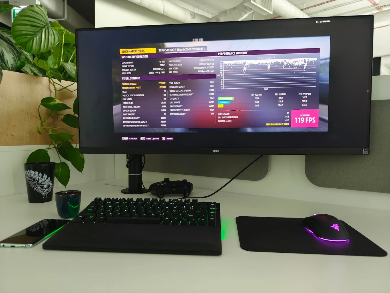 Biurko z dużym monitorem, na którym wyświetlane są dane o wydajności komputera, klawiatura mechaniczna z podświetleniem, mysz oraz telefon. W tle widoczne rośliny doniczkowe.