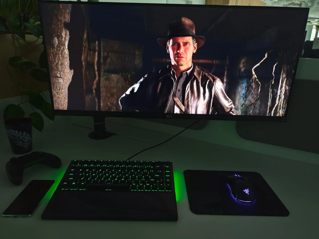 GeForce Now nie narzuca użytkownikom ograniczeń w postaci konieczności ponownego zakupu gier Na ekranie komputera widoczny jest kadr z filmu z mężczyzną w kapeluszu i skórzanej kurtce, przypominający postać z przygodowej serii filmowej. Przed monitorem znajdują się podświetlona klawiatura, mysz komputerowa oraz smartfon na biurku.
