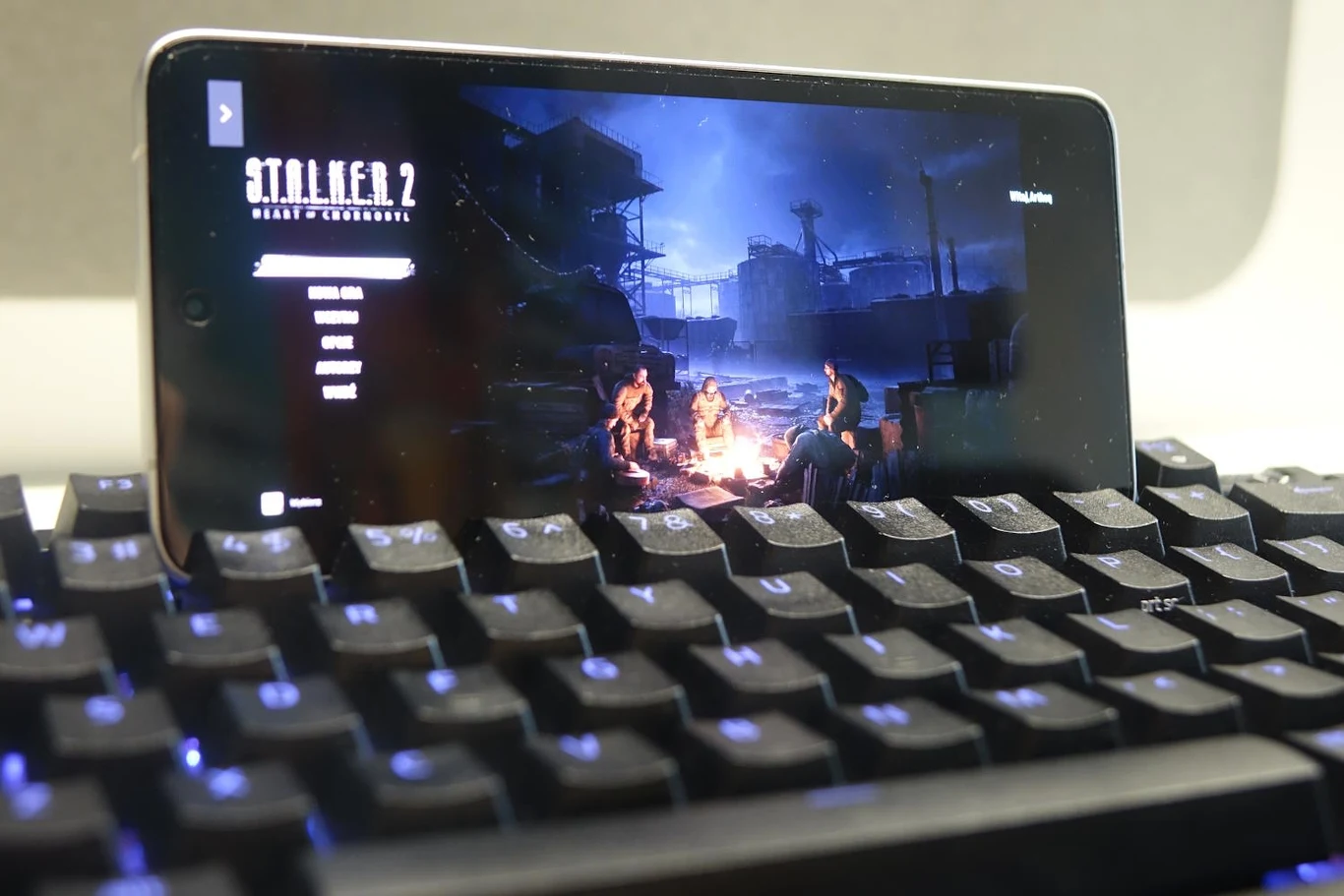 Telefon komórkowy wyświetlający ekran startowy gry Stalker 2 Night of Chernobyl, umieszczony na klawiaturze. Na ekranie widoczna jest scena z postaciami siedzącymi przy ognisku, na tle industrialnego krajobrazu.