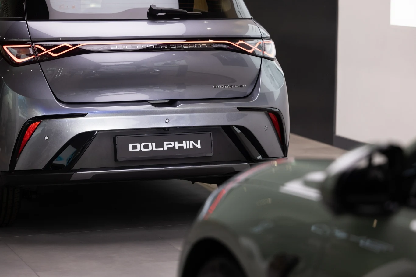 Tylna część szarego elektrycznego samochodu z napisem 'DOLPHIN' na tablicy rejestracyjnej. W tle widać fragment innego pojazdu, częściowo zamazanego i niewyraźnego, ustawionego pod kątem. Wnętrze salonu ma nowoczesny wystrój.