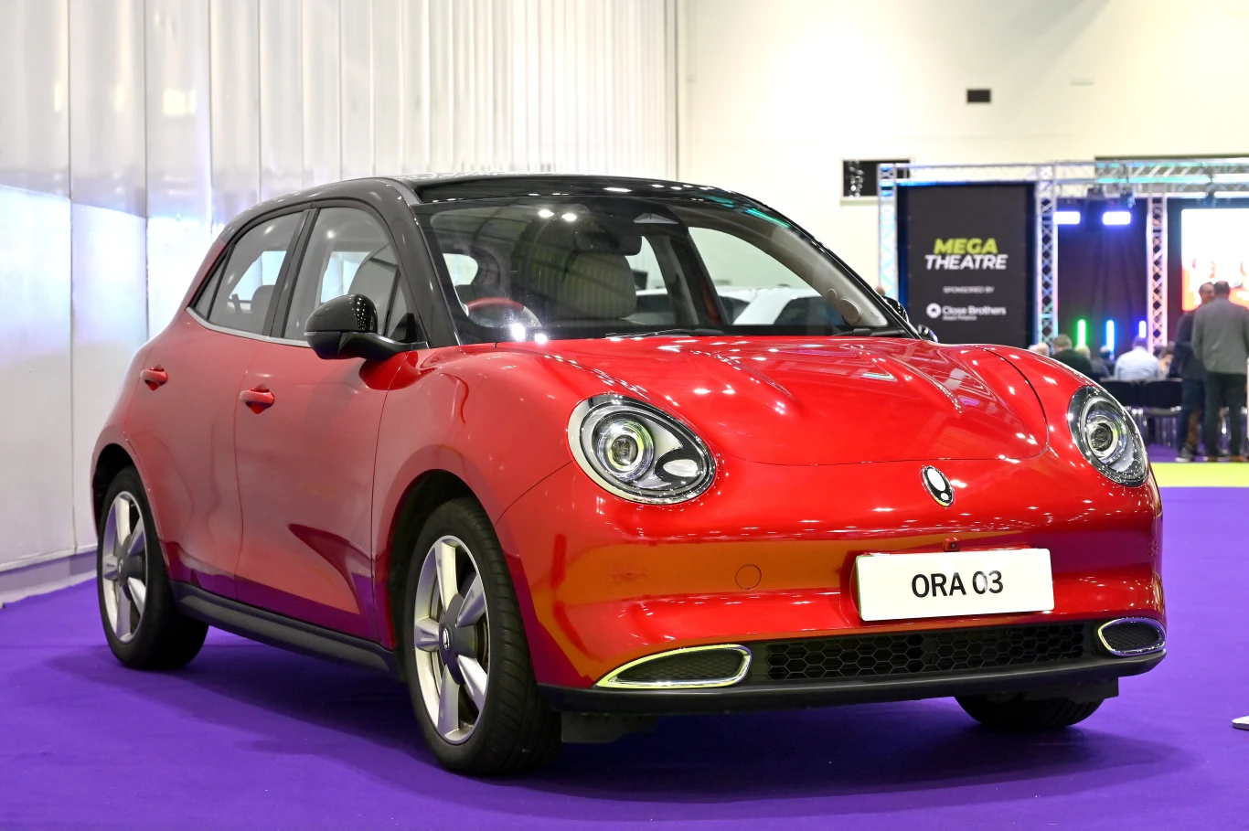 Czerwony samochód ORA O3 prezentowany na podłodze o fioletowym kolorze, w tle banery i inne elementy targowe.