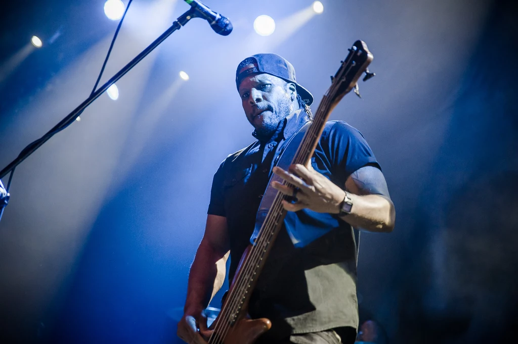 P.O.D. w Warszawie Muzyk grający na gitarze basowej na scenie w trakcie koncertu, oświetlony jasnym światłem reflektorów. Artysta nosi czapkę z daszkiem i skupia się na grze.