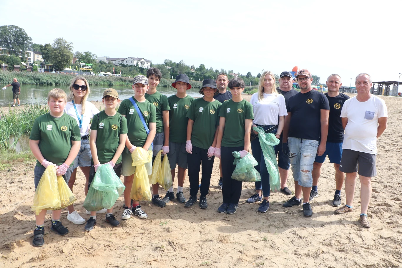 Grupa ludzi stojąca na plaży, ubrana w zielone koszulki, trzymająca worki na śmieci różnego koloru. W tle widoczne jest jezioro z pomostem oraz budynki. Na brzegu widać roślinność.