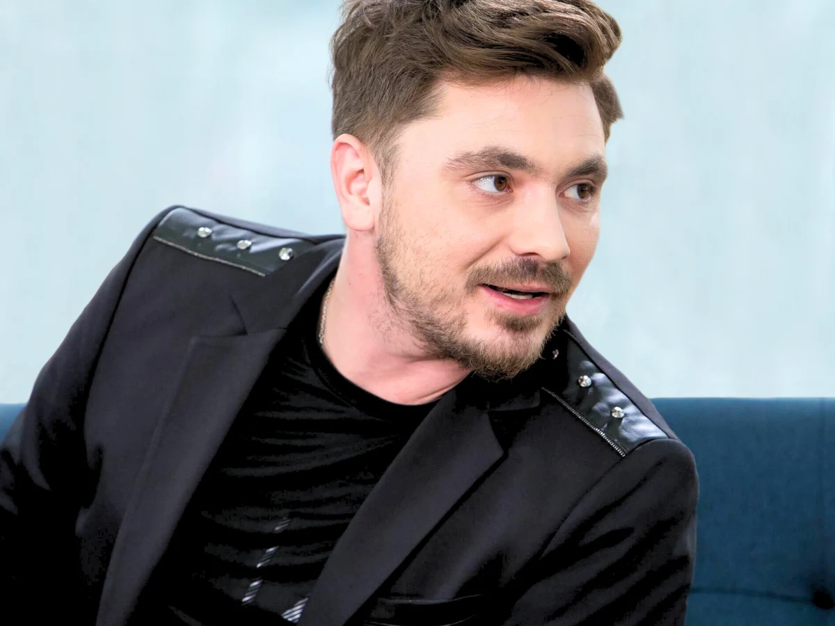 Daniel Martyniuk Daniel Martyniuk z zarostem w ciemnej marynarce z ozdobnymi ramionami, siedzący na niebieskiej sofie, patrzy w bok.