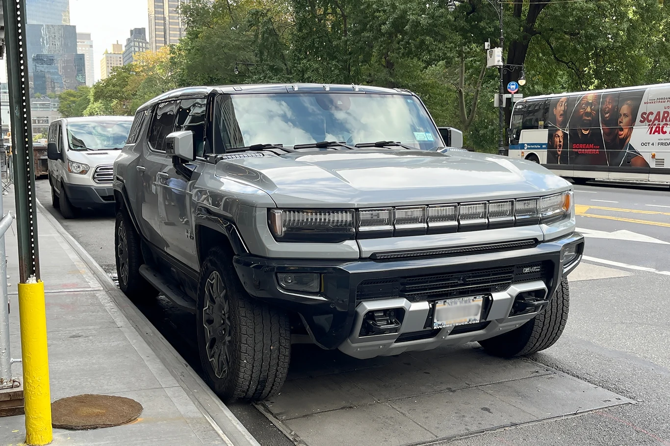 Duże elektryki groźne niczym czołgi. Niszczą zapory drogowe Samochód typu SUV zaparkowany przy chodniku w miejskim otoczeniu. W tle widoczne są drzewa, budynki oraz przejeżdżający autobus.