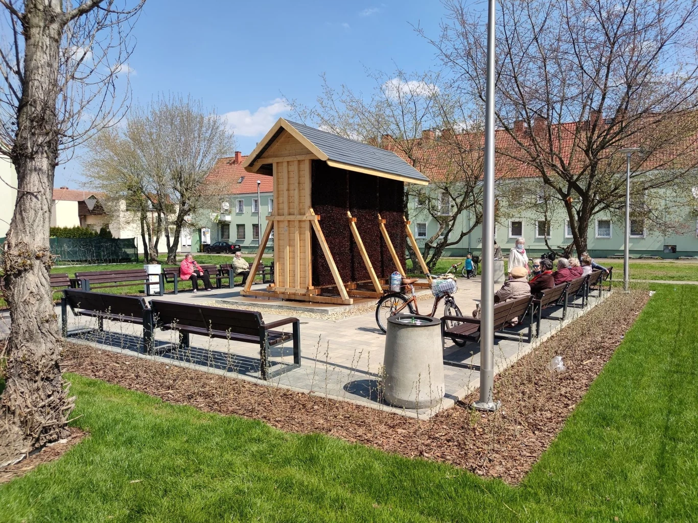 Mały park miejski z drewnianą konstrukcją przypominającą hotel dla owadów w centrum, otoczony ławkami. Kilku ludzi odpoczywa na ławkach, jeden z nich przypiął rower do stojaka. W tle widać niewielkie domy i drzewa.