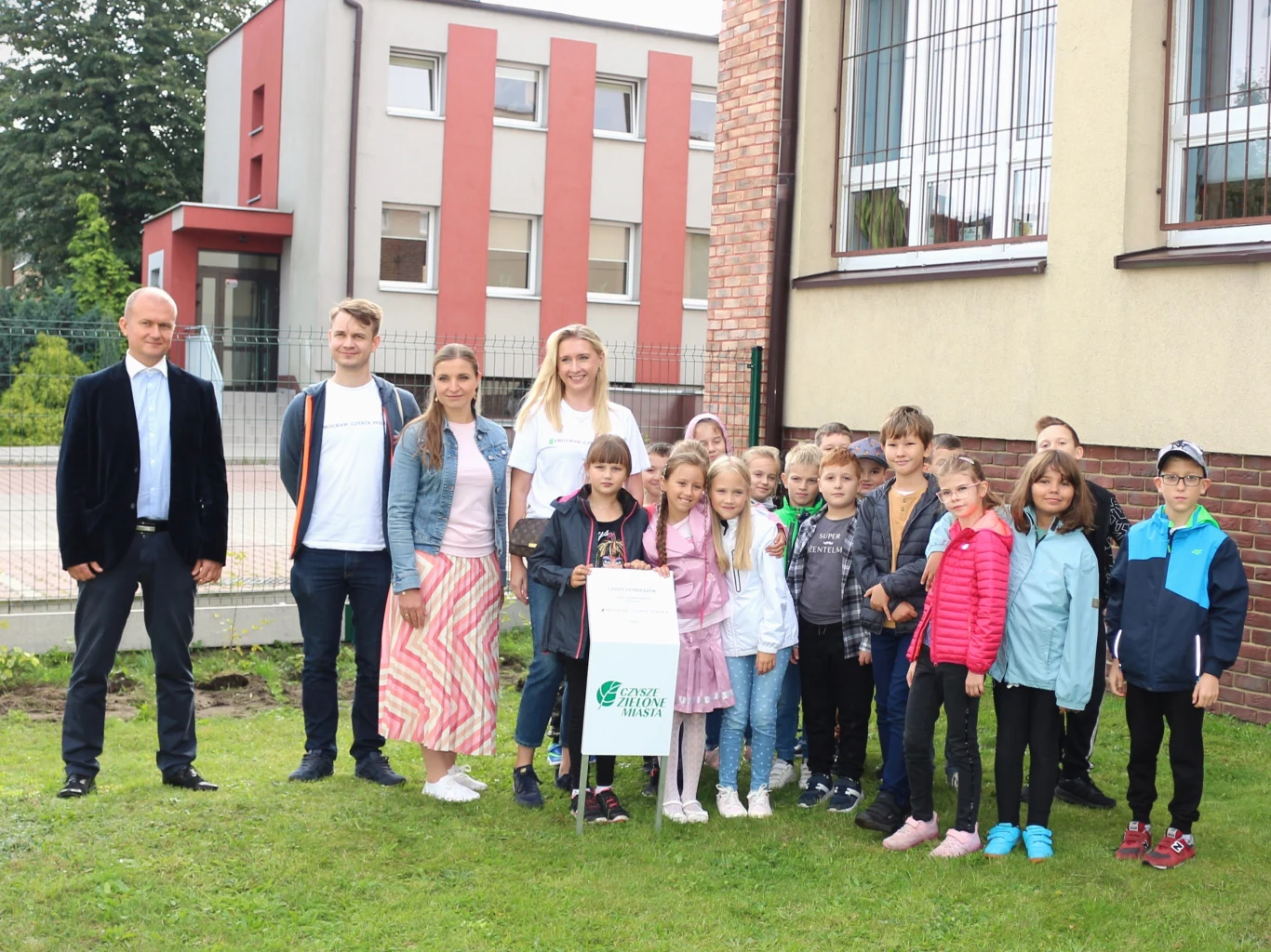 Grupa dzieci stoi na trawie przed budynkiem, z kilkoma dorosłymi osobami po bokach. Dzieci i dorośli są uśmiechnięci. Budynek w tle jest nowoczesny, z dużymi oknami i detalami z czerwonej cegły.