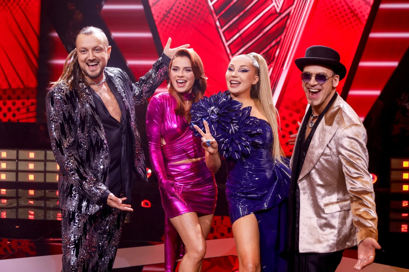 Trenerzy ósmej edycji "The Voice Kids": Baron, Natasza Urbańska, Cleo i Tomson Cztery osoby w eleganckich, kolorowych strojach pozują uśmiechnięte na tle czerwonej scenografii w studiu telewizyjnym. Stroje są błyszczące i nowoczesne.