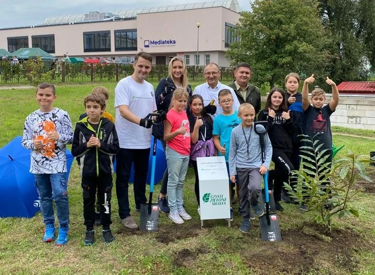 Grupa dzieci i dorosłych pozuje z łopatami na terenie zielonym, przed budynkiem z napisem Mediateka. W tle widoczne są drzewa i budynek. Przed nimi znajduje się młode drzewko, które wygląda jakby zostało niedawno posadzone.