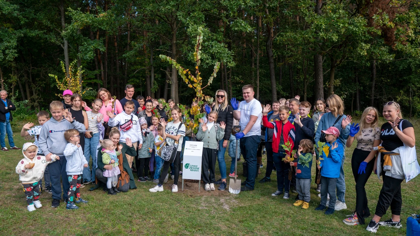 Duża grupa osób, w tym wiele dzieci, pozuje do zdjęcia na tle lasu. W centrum znajduje się tablica informacyjna oraz drzewo. Ludzie wyglądają na radosnych i zadowolonych, niektórzy trzymają liście bądź inne przedmioty związane z naturą.