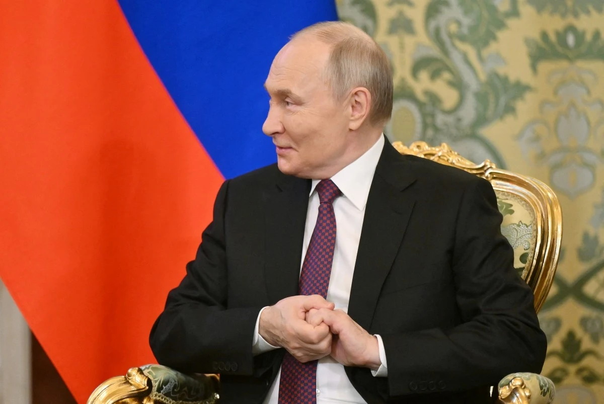 Władimir Putin Mężczyzna w garniturze siedzi na ozdobnym złotym fotelu z rękami splecionymi, w tle widoczne są zasłony w kolorach rosyjskiej flagi.