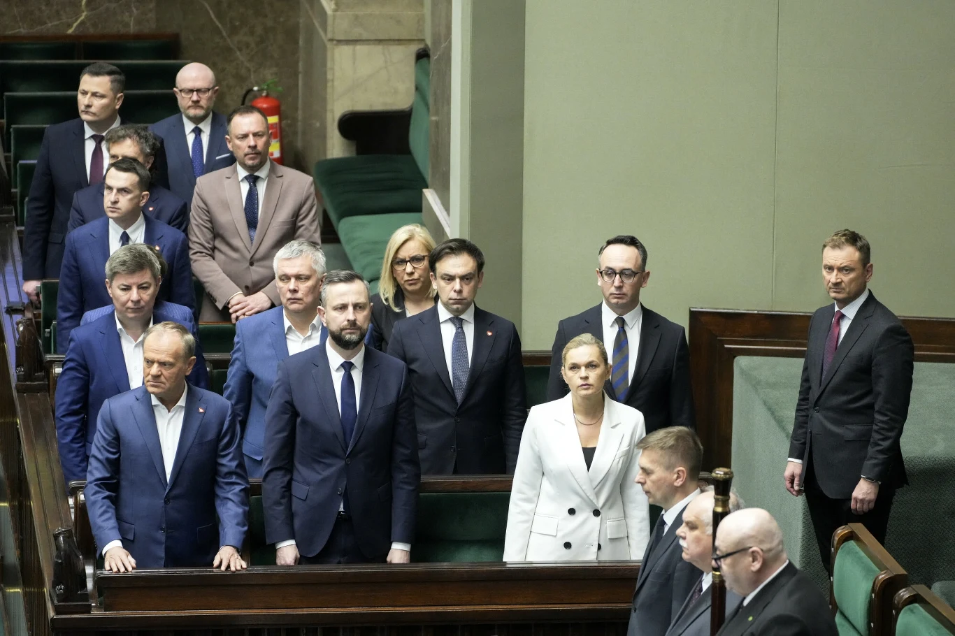 Sondaż CBOS. Polacy oceniają rząd Donalda Tuska Politycy w sali obrad, ubrani w formalne garnitury oraz sukienki, stoją w ławach poselskich podczas sesji parlamentarnej. Widać poważne wyrazy twarzy. Pomieszczenie jest oficjalne i eleganckie.