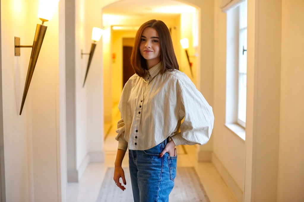 Alicja Wieniawa-Narkiewicz Młoda kobieta w luźnej, eleganckiej koszuli i jeansach stoi w dobrze oświetlonym, stylowym wnętrzu, uśmiechnięta. W tle jasne lampy i korytarz dodają scenie przytulnego charakteru.