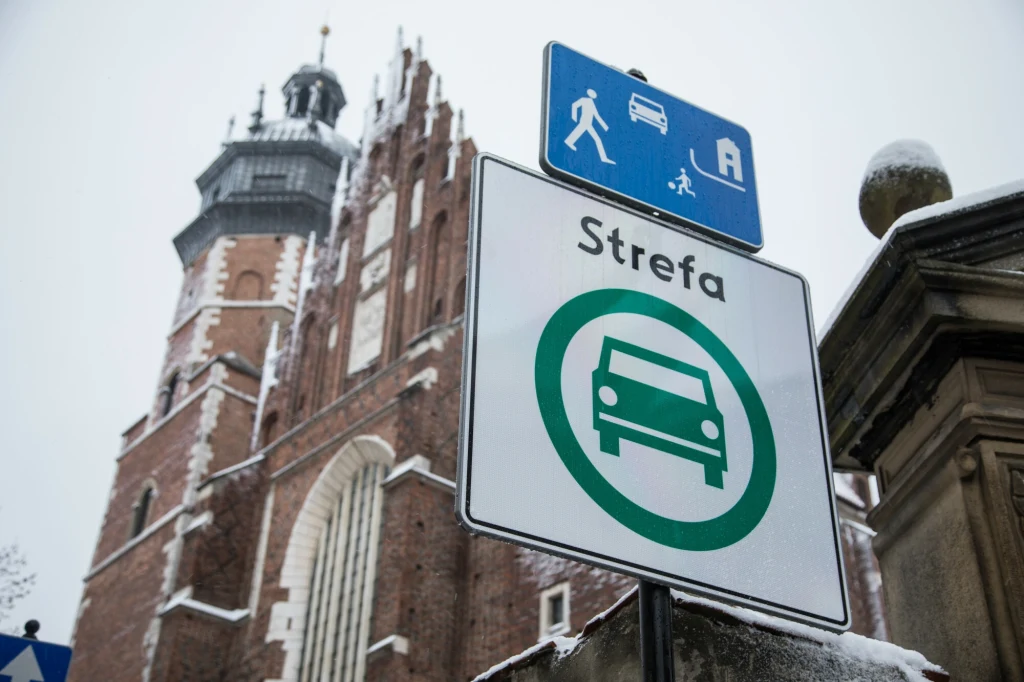 Od stycznia rusza SCT w Krakowie. Jak miasto będzie ją kontrolować? Znak drogowy z symbolem strefy i samochodem na tle gotyckiego kościoła, możliwe że to Kościół Mariacki w Krakowie.