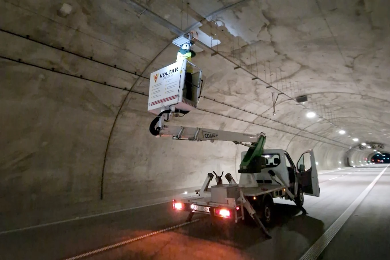 Pracownik w koszu podnośnika wykonywujący prace konserwacyjne na suficie tunelu. Samochód z wysięgnikiem zaparkowany jest na pasie ruchu, wokół widoczne są światła tunelu.