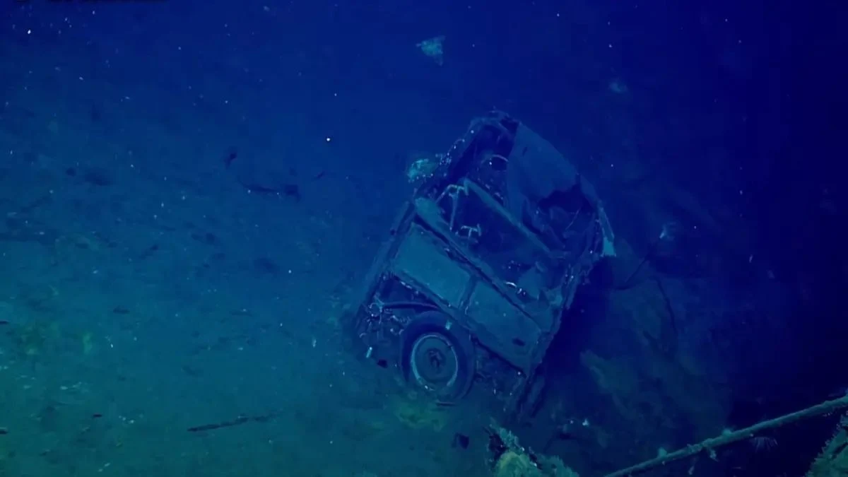 Częściowo zachowany pojazd osobowy odkryty na USS Yorktown, lotniskowcu zatopionym przez japońskie siły podczas II wojny światowej Wrak pojazdu spoczywa na dnie morskim, otoczony mglistym, niebieskawym tłem. Widoczne są niektóre szczegóły konstrukcji, takie jak koła i rama, a wokół widać osady i drobne organizmy morskie.