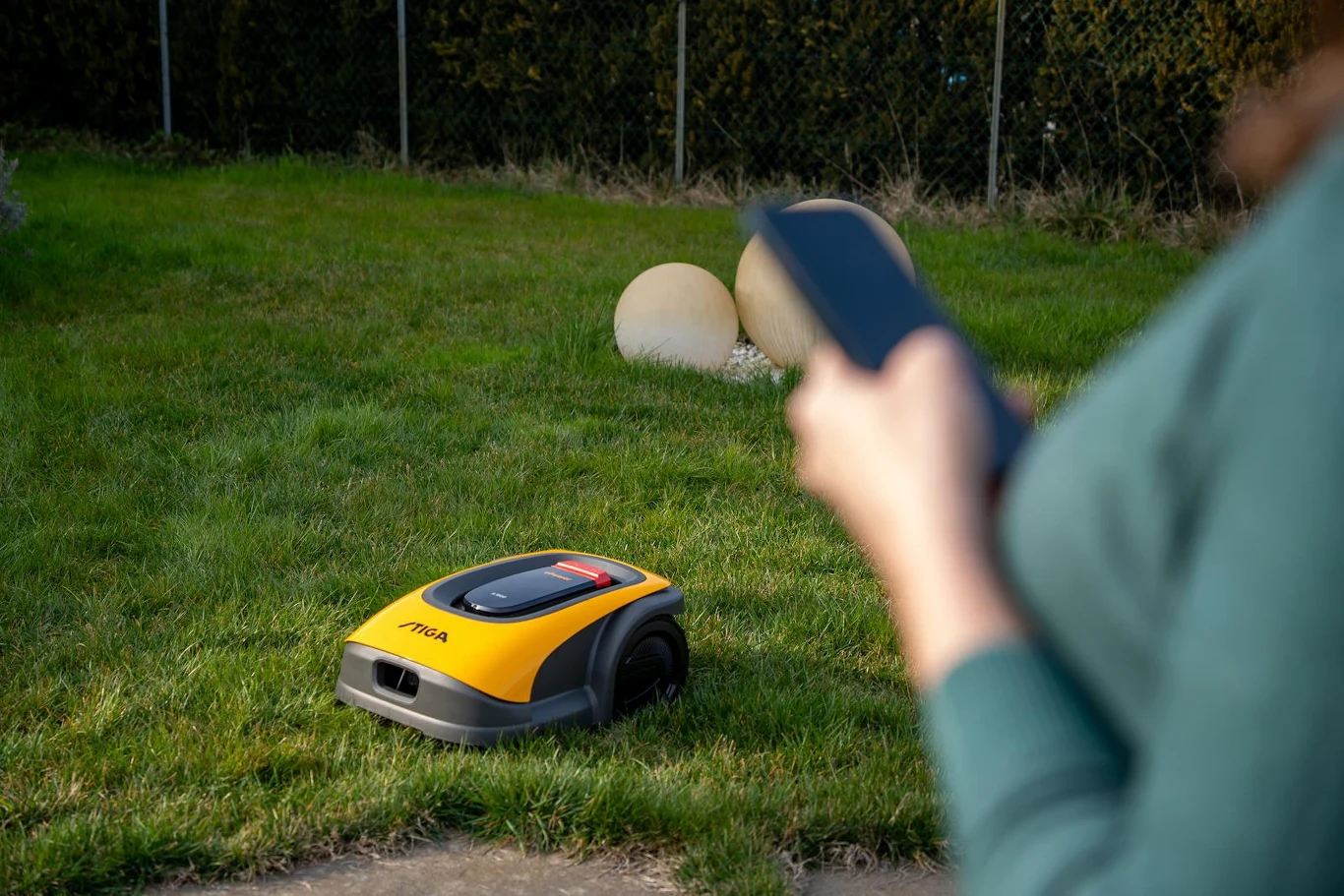 Niektórzy traktują koszenie jako relaks, inni wolą zautomatyzować ten proces Automatyczna kosiarka poruszająca się po trawniku, sterowana przez osobę trzymającą urządzenie mobilne. W tle widoczne są kamienne dekoracje ogrodowe.