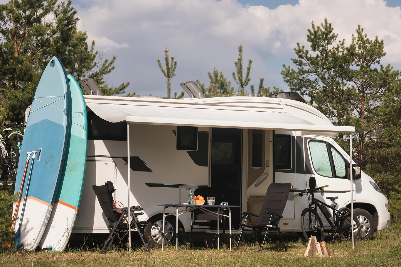Rusza sezon karawaningowy. Nie popełnij podstawowych błędów Kamper zaparkowany na trawiastej polanie, obok którego stoi deska surfingowa, rower i składany stolik z krzesłami. W tle widoczne sosnowe drzewa i błękitne niebo z chmurami. Atmosfera wypoczynku i rekreacji na świeżym powietrzu.