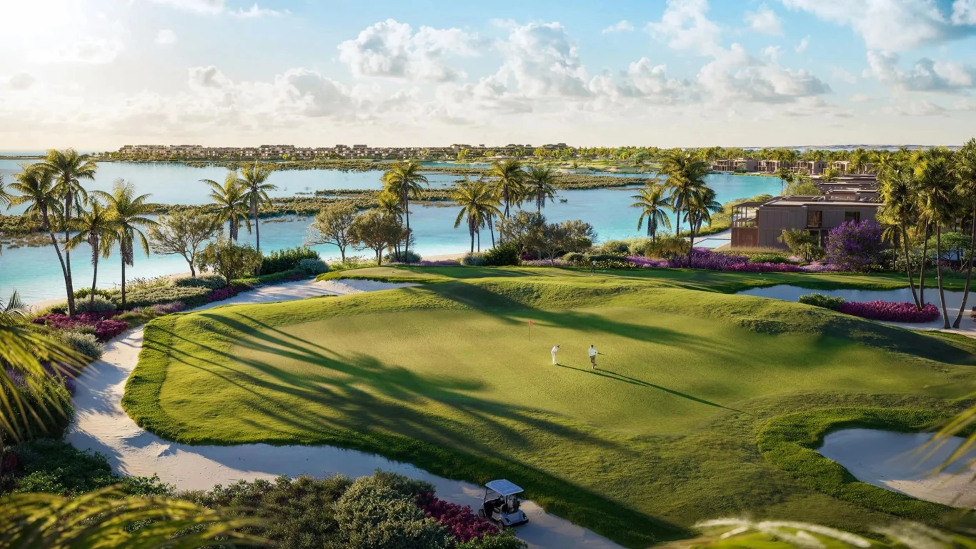 Zielone pole golfowe otoczone palmami i kolorowymi kwiatami, z widokiem na malowniczy krajobraz przybrzeżny. W tle błękitne niebo z białymi chmurami oraz spokojna woda jeziora. Na polu znajdują się dwie osoby i wózek golfowy.