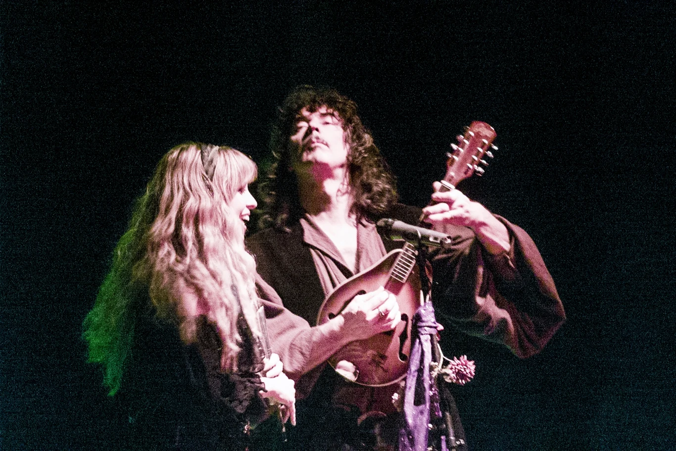 Dwóch muzyków na scenie wykonujących utwór, jeden gra na gitarze akustycznej, drugi trzymał mandolinę. Atmosfera jest pełna pasji i energii, oświetlenie sceniczne podkreśla emocje towarzyszące występowi.