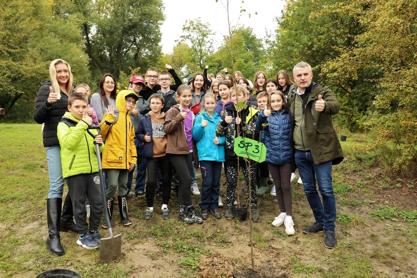 Grupa dzieci i dorosłych stoi razem w parku podczas sadzenia drzewa. Wszyscy są uśmiechnięci i pokazują gesty aprobaty. Na pierwszym planie znajduje się też symboliczne drzewo z tabliczką oznaczoną 'SP3'.