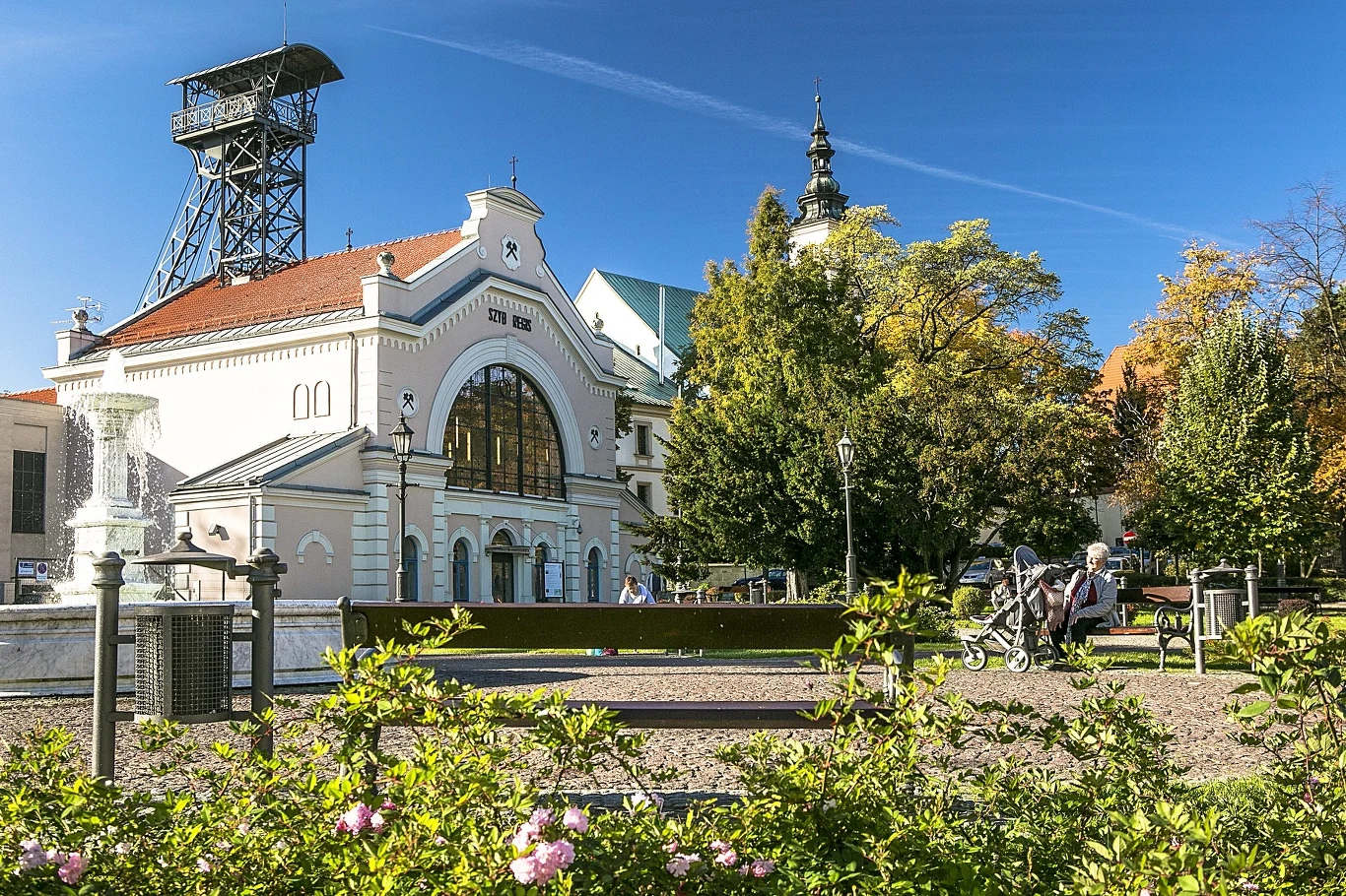 Zabytkowy budynek kopalni w otoczeniu zieleni, z charakterystyczną wieżą szybów górniczych, otoczony przez alejki spacerowe i drzewa w jesiennych kolorach. W tle niebo bez chmur.