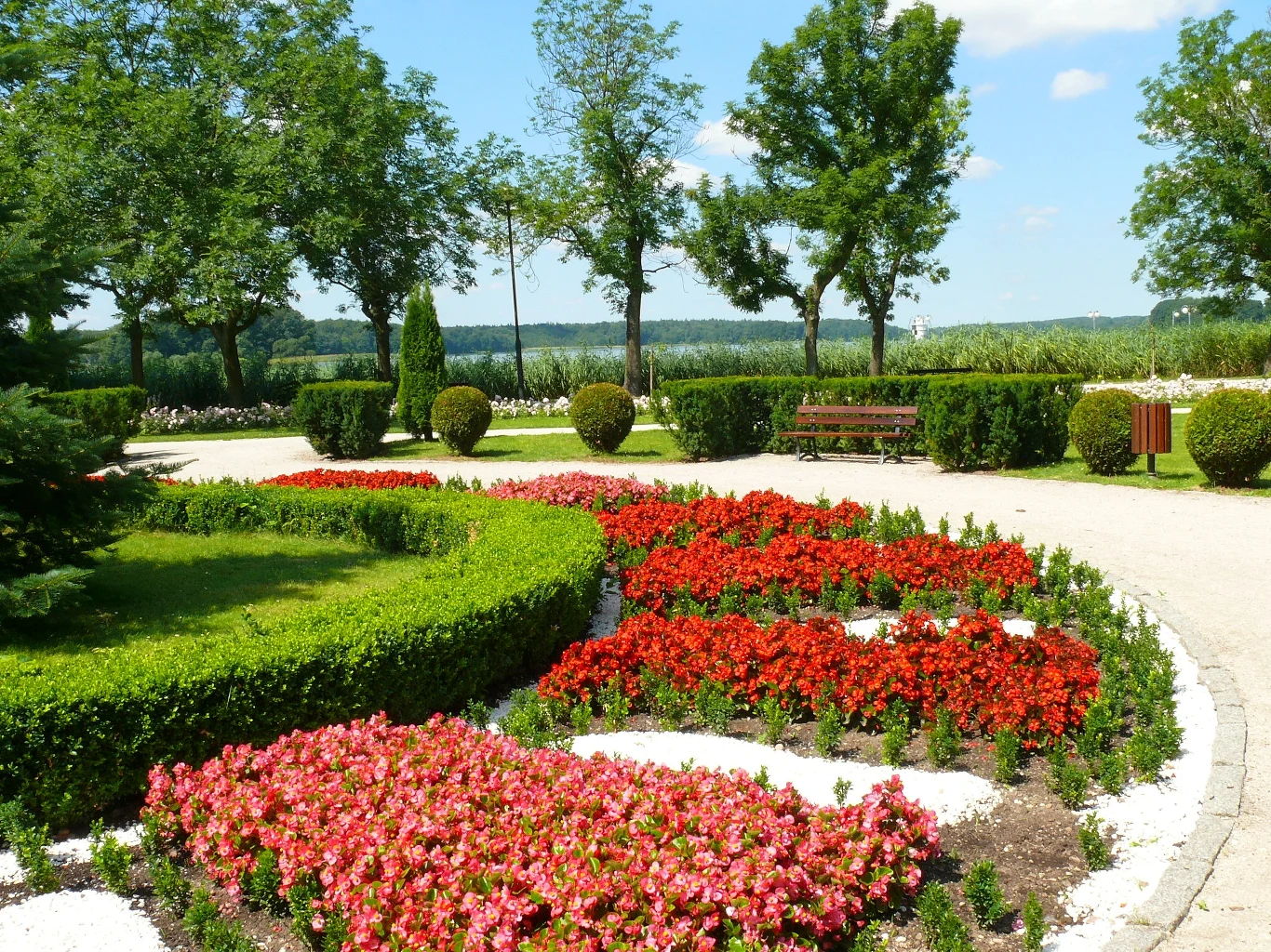 Zadbany ogród z różnokolorowymi kwiatami w foncie, otoczony drzewami i żywopłotem, w tle widać alejki i ławkę. Piękne otoczenie tworzy atmosferę spokoju i relaksu, sprzyja odpoczynkowi na świeżym powietrzu.