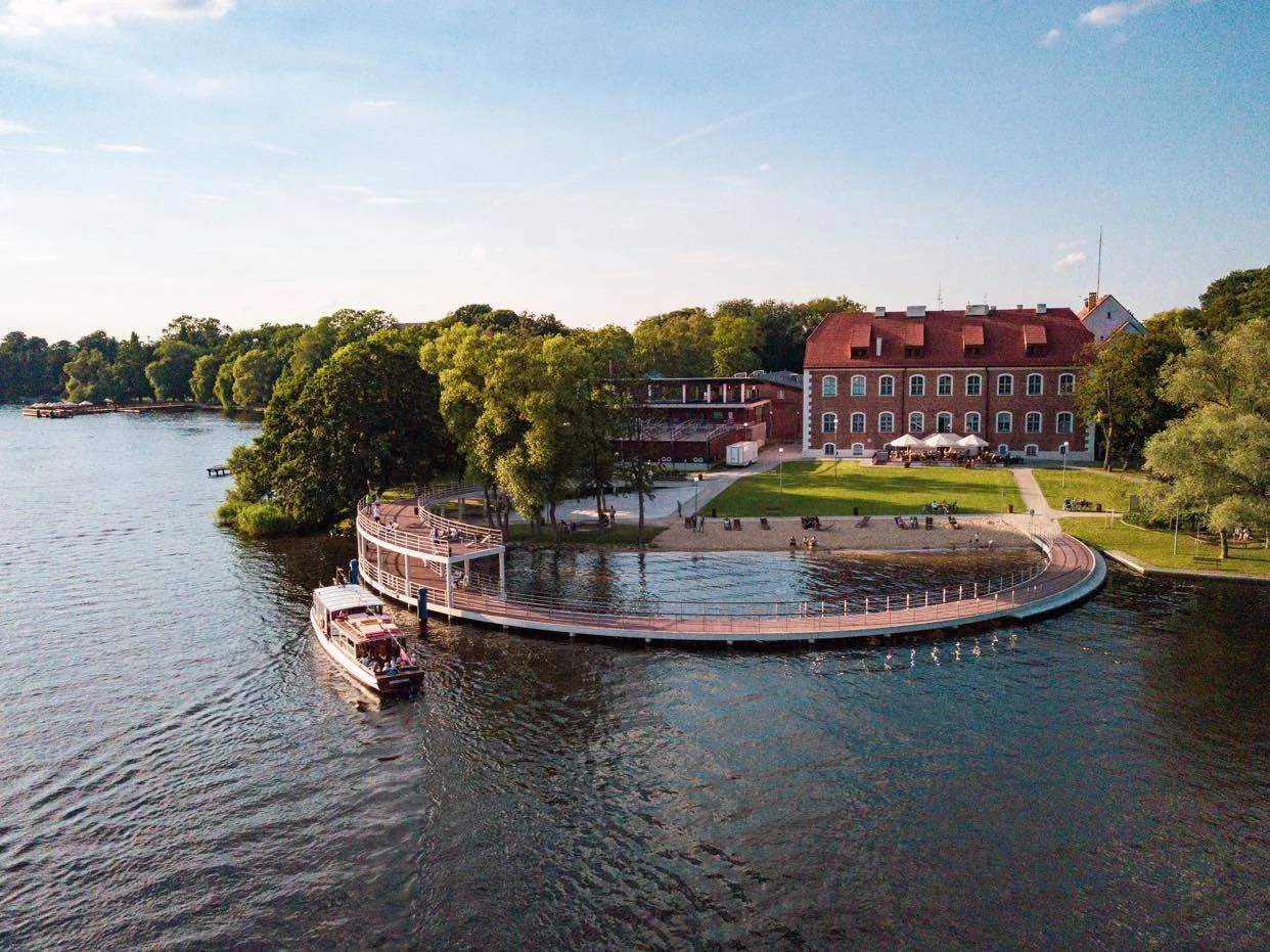 Widok na przystań nad jeziorem z budynkiem z czerwonym dachem po prawej stronie. Na wodzie pływa statek turystyczny, a całość otoczona jest zielonymi drzewami i parkiem.