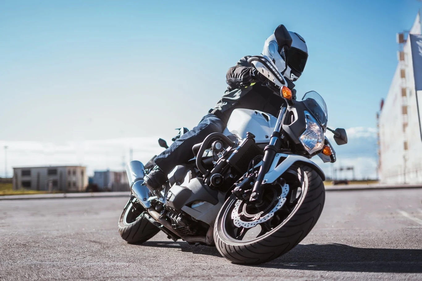 Motocyklista ubrany w pełny strój ochronny pochyla się na zakręcie, jadąc nowoczesnym motocyklem po pustej drodze. W tle niebo z nielicznymi chmurami i zarys budynków.