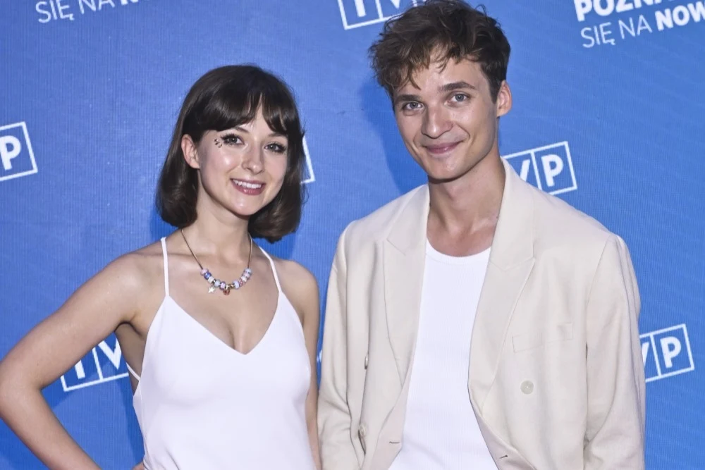 Kasia i Jacek Sienkiewiczowie z zespołu Kwiat Jabłoni Mężczyzna i kobieta na niebieskim tle z logotypami, oboje ubrani w białe ubrania, uśmiechnięci.