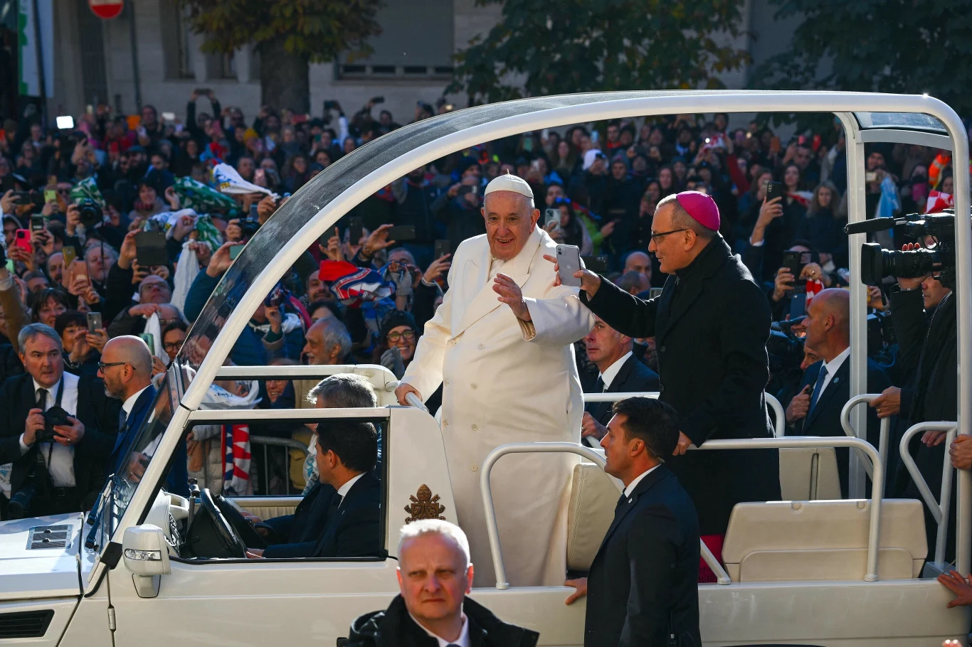 Papież w otwartym samochodzie papamobile pozdrawia tłum zgromadzony na ulicy. Obok niego stoi duchowny, a w tle widać wielu ludzi machających i robiących zdjęcia. Uroczysta atmosfera i silna ochrona wokół pojazdu.