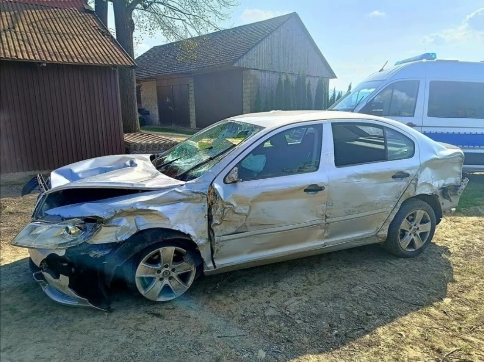 10-latek zginął pod kołami skody, którą prowadził pijany 23-latek (mat. prasowy-edytowany) Zniszczony samochód osobowy stoi na wiejskiej drodze obok ceglanych budynków. Auto ma zgnieciony przód oraz zarysowane boki. W tle widoczny jest radiowóz policyjny.