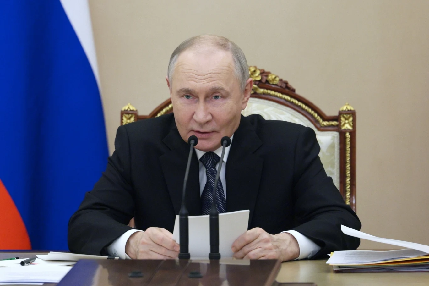 Prezydent Rosji Władimir Putin Mężczyzna w garniturze przemawia do mikrofonów, siedząc przy stole. Na stole znajdują się dokumenty. W tle widoczne są fragmenty flagi.