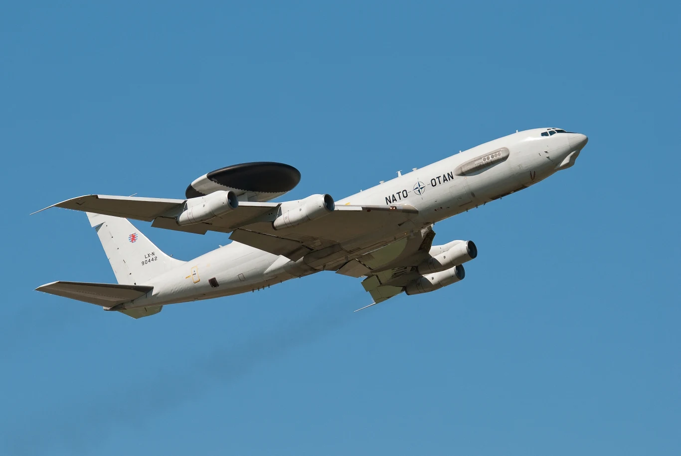 Samolot NATO z systemem wczesnego ostrzegania i kontroli AWACS w locie na tle błękitnego nieba, widoczna charakterystyczna tarcza na górze samolotu, symbol organizacji na kadłubie.