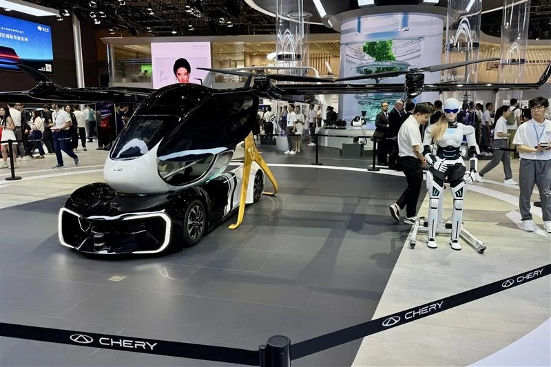 Elektryczny pojazd latający z rotorami w hali wystawowej, otoczony przez odwiedzających. Obok pojazdu stoi humanoidalny robot, a na podłodze widoczny jest znak marki Chery.