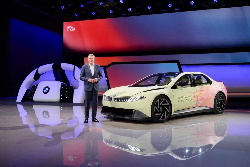 Mężczyzna prezentujący nowy model futurystycznego samochodu elektrycznego na tle ekranu z logo marki i modernistycznym otoczeniem.