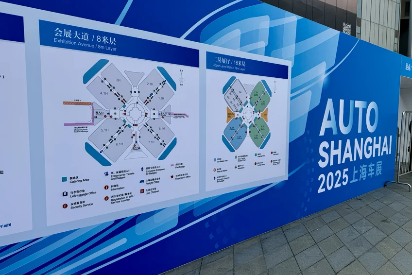 Plan wystawowy na dużym plakacie z napisem 'Auto Shanghai 2025' na niebieskim tle, przedstawiający schemat rozmieszczenia stoisk na wydarzeniu motoryzacyjnym.