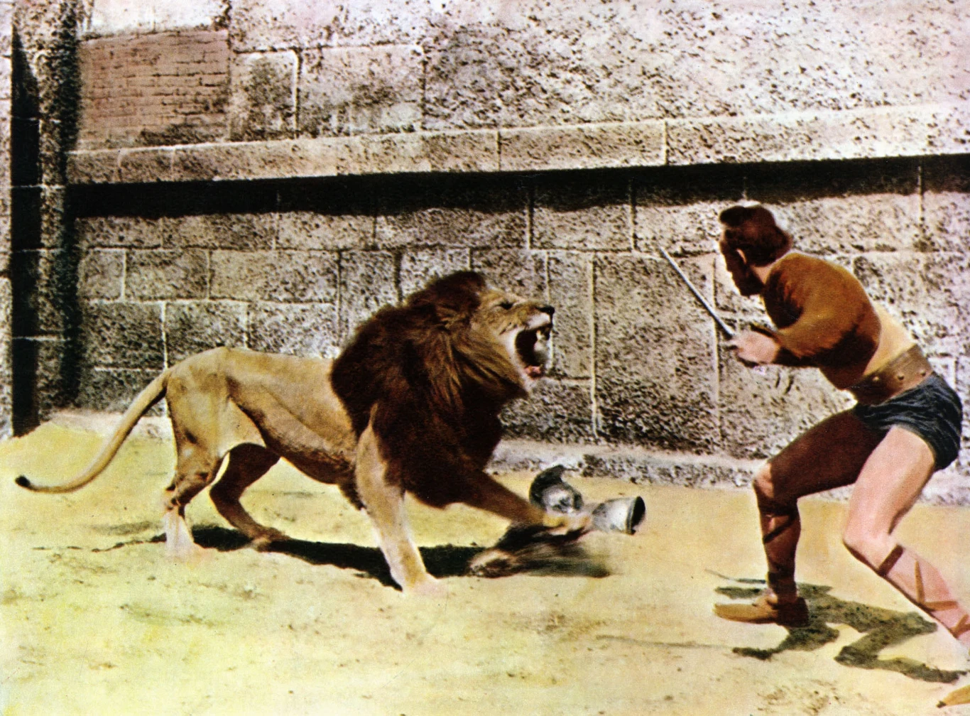 Człowiek trzymający sztylet stawia czoła rozwścieczonemu lwu na arenie otoczonej kamiennym murem, scena przypomina walki gladiatorów z czasów starożytnych Rzymian.
